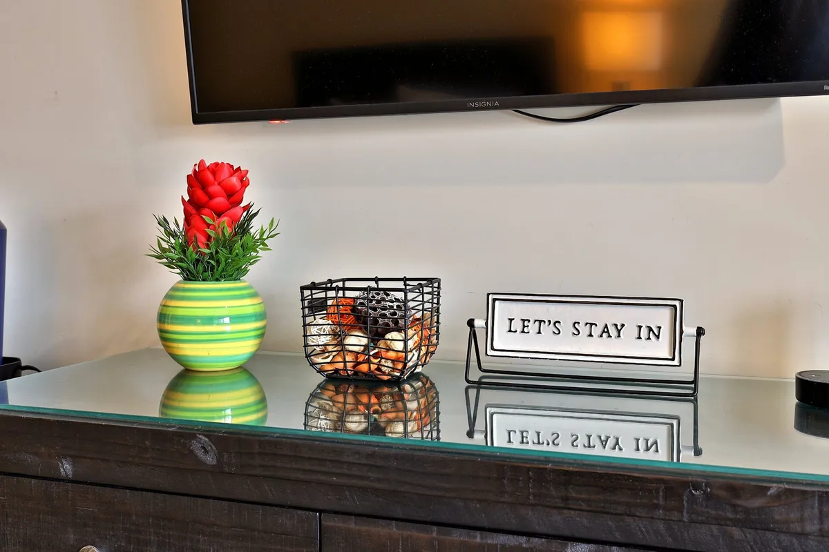 A welcoming interior scene featuring a vibrant red flower arrangement in a striped green vase, a decorative basket with shells, and a motivational sign. The ambiance suggests a cozy space ideal for relaxing indoors, sharing light conversation, or unwinding after a day out.