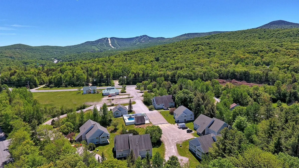 An exterior view showing mountains in the distance, a cluster of buildings nestled among greenery, and a swimming pool in the foreground. The landscape highlights the property's proximity to natural beauty and recreational amenities.