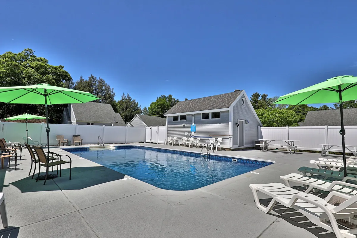 A cheerful scene of the swimming pool area with green umbrellas, plenty of lounge chairs, and mature trees in the background. The late daytime light suggests relaxed, extended hours for gathering and enjoying summer evenings by the pool.