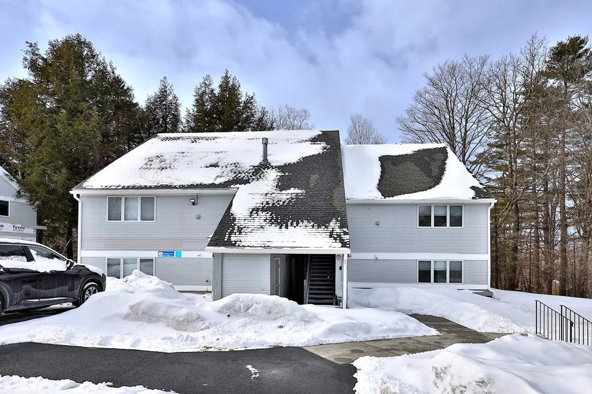 A snow-covered building exterior with entrance stairs, surrounded by tall trees. The scene evokes a serene winter atmosphere, capturing the feeling of arriving at a cozy mountain retreat just moments from the slopes.