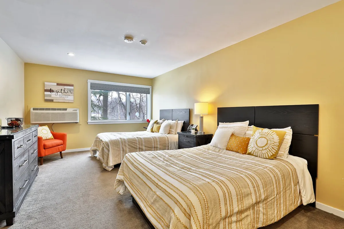 A bright studio space featuring two beds positioned near a large window, a dresser, and an accent chair. Morning sunlight pours into the room, giving it a warm and inviting feel. The arrangement encourages relaxation and social interaction after a day on the slopes or trails.