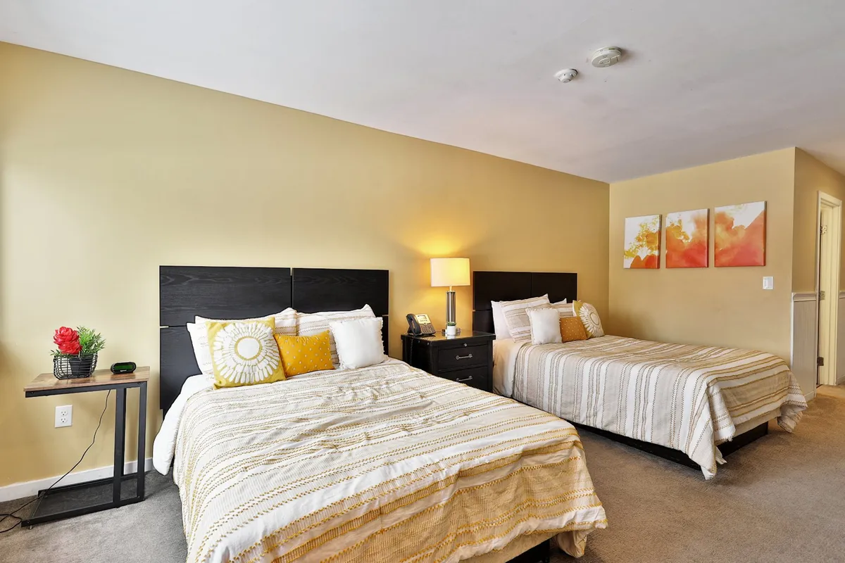 A soothing sleeping area with two beds, a bedside lamp, tasteful artwork, and a nightstand adorned with a plant. The space feels tranquil and is styled with modern, calming décor, emphasizing a restful retreat.