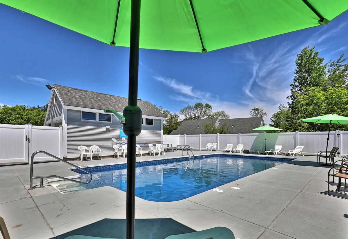 The image features the complex's swimming pool bordered by green umbrellas and lounge chairs, with ample space for guests to relax or socialize. The lighting and composition evoke the feeling of a lively but laid-back summer gathering.